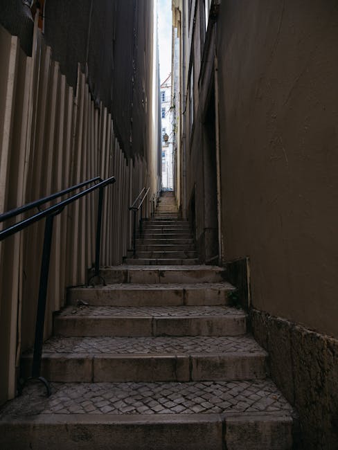 A narrow outdoor staircase with stone steps leading upward between two tall, enclosed walls—one on the left with a black metal railing and a temporary construction barrier, and one on the right with a smooth, painted surface. The steps are slightly weathered and uneven. At the top of the staircase, there is a view of a narrow street with buildings visible in the distance. The staircase is part of a home relocation process, illustrating the constrained space often encountered during furniture transport in urban areas, as handled by Fulham Removals, a professional removals company specializing in tight staircase access.
