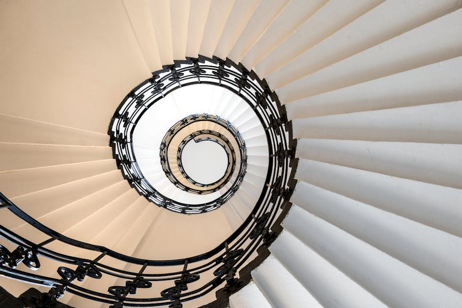 A view from the top of a spiral staircase showing a circular iron railing painted black, with beige carpeted stairs curving downwards in a tight spiral. The stairs are illuminated by natural light from above, creating a bright contrast with the dark railing. The image captures the intricate design of the staircase, commonly encountered during home relocations where precise furniture transport and packing are necessary to navigate tight staircases. Fulham Removals, specialists in removals services, often assist with such challenging moving logistics, including the careful loading and unloading of furniture and boxes from properties featuring spiral staircases, as shown here.