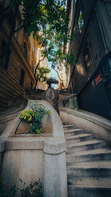 A narrow outdoor staircase with stone steps leading upward between two tall, enclosed walls—one on the left with a black metal railing and a temporary construction barrier, and one on the right with a smooth, painted surface. The steps are slightly weathered and uneven. At the top of the staircase, there is a view of a narrow street with buildings visible in the distance. The staircase is part of a home relocation process, illustrating the constrained space often encountered during furniture transport in urban areas, as handled by Fulham Removals, a professional removals company specializing in tight staircase access.
