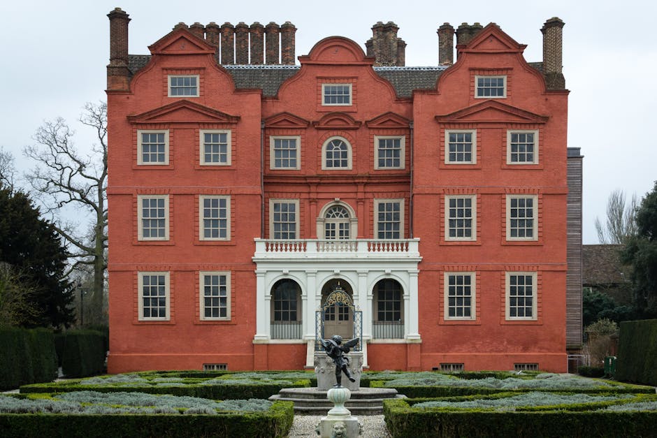 A large, historic red brick house with a symmetrical façade, featuring three stories and multiple evenly spaced rectangular windows with white frames. The central entrance has a white portico supported by columns and a decorative balcony above. In front of the building, there is a neatly maintained gravel pathway leading to the entrance, bordered by well-trimmed hedges and a symmetrical garden layout. A stone fountain with a sculpted figure is positioned on the pathway, with water not visible. The scene is outdoors during daytime with an overcast sky, and nearby trees and bushes are visible to the sides of the property. The image supports themes of home relocation and furniture transport, relevant to Fulham Removals' house removals service, especially in the context of moving historic homes, with elements like the building’s structure and garden emphasizing the importance of careful packing and logistical planning during a move.