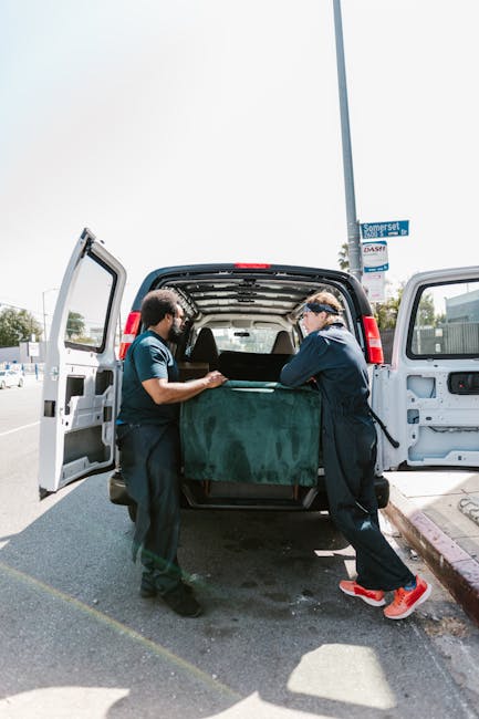 Two men wearing dark work uniforms and gloves are engaged in a home relocation task, standing outside a white cargo van with its rear doors open. They are lifting a large, rectangular piece of furniture or packaging wrapped in dark green fabric or protective material into the van, which is parked on a street with a clear sky overhead. The interior of the van shows a spacious loading area with a black carpeted floor, and the men are using their hands to carefully handle the item, suggesting a careful loading process. In the background, there are street signs, a lamppost, and some buildings, indicating an urban environment suitable for furniture transport and packing during house removals. This scene illustrates the practical steps involved in furniture transport and packing during house removals, as managed by professional services like Fulham Removals, supporting efficient home relocation processes for historic homes and modern residences alike.