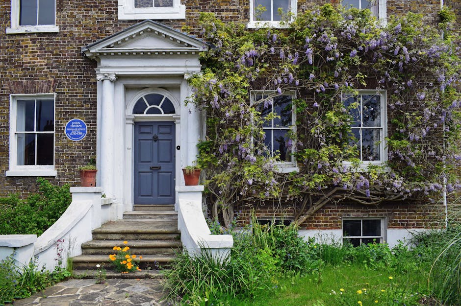A large, historic red brick house with a symmetrical façade, featuring three stories and multiple evenly spaced rectangular windows with white frames. The central entrance has a white portico supported by columns and a decorative balcony above. In front of the building, there is a neatly maintained gravel pathway leading to the entrance, bordered by well-trimmed hedges and a symmetrical garden layout. A stone fountain with a sculpted figure is positioned on the pathway, with water not visible. The scene is outdoors during daytime with an overcast sky, and nearby trees and bushes are visible to the sides of the property. The image supports themes of home relocation and furniture transport, relevant to Fulham Removals' house removals service, especially in the context of moving historic homes, with elements like the building’s structure and garden emphasizing the importance of careful packing and logistical planning during a move.