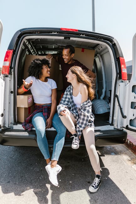 Two young women sit inside the open back of a moving van parked on a pavement, engaged in conversation and smiling. They are surrounded by packed cardboard boxes and protective wrapping materials, indicating a home relocation or packing process. One woman has curly hair and wears a white T-shirt and jeans, while the other has straight hair and wears a sleeveless top with a plaid shirt tied around her waist and light-colored leggings. A man stands inside the van, leaning into the cargo area and smiling at them. The interior of the van shows additional packing supplies and furniture wrapped for transport. The scene captures a moment of loading or unloading objects during a move, with bright natural lighting and an urban setting, aligning with professional removals and relocation activities typical of house move logistics. Fulham Removals offers experienced furniture transport and packing services, essential for navigating narrow roads and parking restrictions in locations like Parsons Green.
